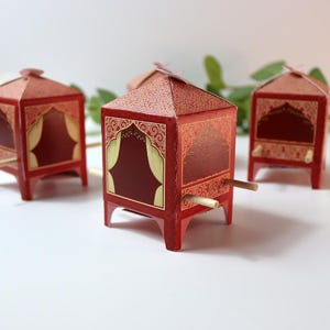 Puede incluir: Cajas decorativas de papel rojo con detalles dorados y patrones intrincados. Las cajas tienen una forma &uacute;nica, que se asemeja a peque&ntilde;as estructuras con aberturas arqueadas y clavijas de madera. El dise&ntilde;o incorpora una rica paleta de colores y detalles ornamentados.