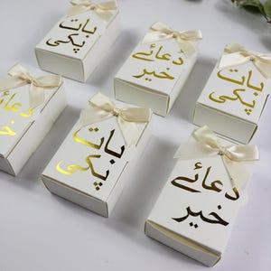May include: A collection of rectangular white gift boxes, each adorned with a cream-colored satin ribbon bow. The boxes feature gold foil calligraphy text in Urdu, with some boxes displaying the phrase Baat Pakki and others showing Dua-e-Khair.