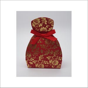 Puede incluir: Una pequeña caja de regalo curva con una base de color rojo intenso y un patrón floral dorado repetitivo. La parte superior de la caja está fruncida y asegurada con una cinta de tela roja brillante atada en un lazo, creando una silueta redondeada.