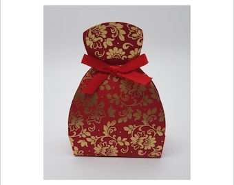 May include: A small, curved gift box featuring a deep red base color with a repeating gold floral pattern. The top of the box is cinched and secured with a bright red fabric ribbon tied into a bow, creating a distinct, rounded silhouette.
