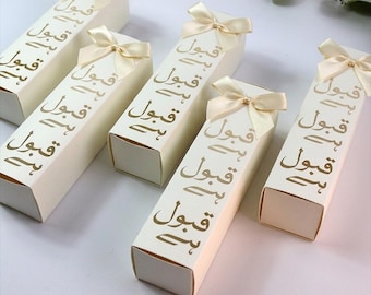 May include: Several cream-colored rectangular gift boxes with gold Arabic script and a cream-colored bow. The boxes are arranged on a white surface. The boxes are closed and ready to be gifted.