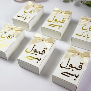 May include: A collection of rectangular white gift boxes, each topped with a cream-colored satin ribbon bow. The boxes feature gold foil calligraphy text in Arabic script, with some reading Nikah Mubarak and others reading Qubool Hai.
