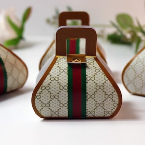 May include: A set of decorative gift boxes shaped like handbags. The boxes feature a repeating infinity symbol pattern on a cream background, with a vertical red and green stripe. Each box has a brown handle and trim.