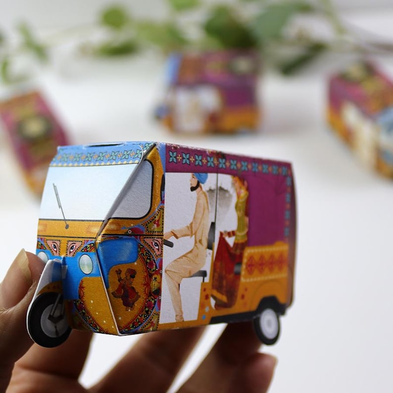 May include: A small paper model of a three-wheeled auto rickshaw featuring vibrant yellow, blue, and purple colors. The side panel displays an illustration of a person driving and a passenger sitting inside, decorated with intricate patterns.