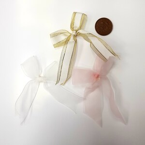 May include: Three decorative bows in white, gold, and pink. The gold bow has a striped design. A penny is next to the bows for size comparison. The bows are made of ribbon and are likely for gift wrapping or crafts.
