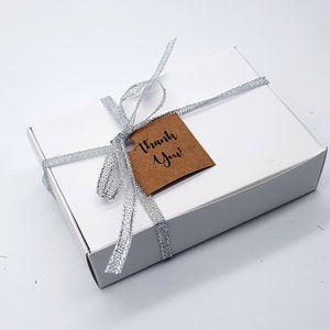 May include: A white rectangular gift box tied with a silver ribbon. A small brown tag with the words "Thank You!" is attached to the ribbon. The box is on a white background.