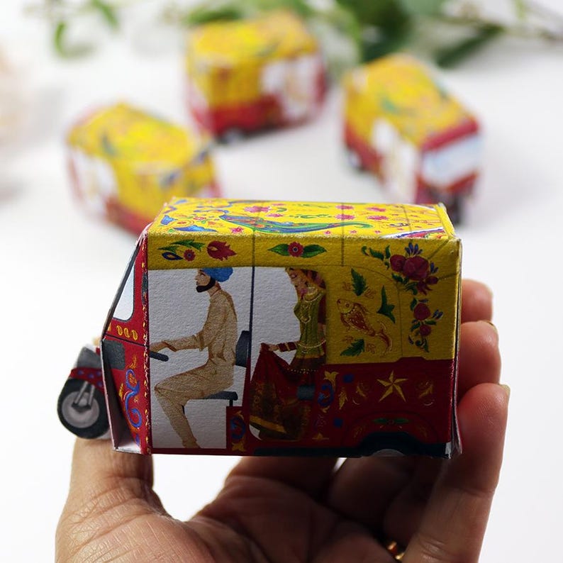 K&ouml;nnte beinhalten: Ein farbenfrohes Miniatur-Papier-Auto-Rikscha mit aufwendigen floralen und fig&uuml;rlichen Illustrationen. Das Fahrzeug hat ein gelbes Dach, rote Akzente und eine detaillierte Darstellung eines Fahrers und eines Passagiers. Das Design erinnert an traditionelle indische Kunst.