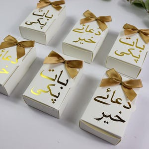 May include: White gift boxes with gold text and gold bows. The rectangular boxes feature gold script text and are each tied with a gold satin bow. The boxes are arranged on a white surface.