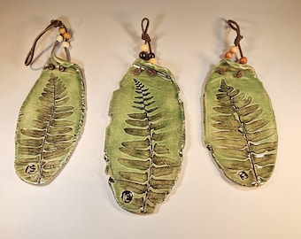 May include: Three oval-shaped ceramic ornaments in a light green glaze, each featuring a dark brown fern leaf imprint. Each piece includes a brown leather cord loop with small wooden beads for hanging and a small stamped maker mark.