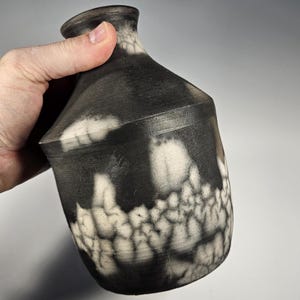 May include: A handmade ceramic vase with a unique black and white design. The vase has a dark, matte finish with a textured, crackled pattern in white. The vase is held by a hand, showcasing its shape and size.