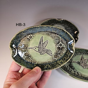 May include: Oval ceramic dish with a hummingbird design. The dish features a light green center with a black hummingbird illustration. The rim is decorated with a lace pattern in shades of blue and green. The text "HB-3" is visible.