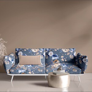 May include: A floral patterned sofa with a white frame and a beige pillow sits in front of a beige wall. A silver round coffee table sits in front of the sofa. A white floor lamp with a beige lampshade is in the corner of the room.