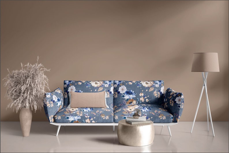 May include: A floral patterned sofa with a white frame and a beige pillow sits in front of a beige wall. A silver round coffee table sits in front of the sofa. A white floor lamp with a beige lampshade is in the corner of the room.