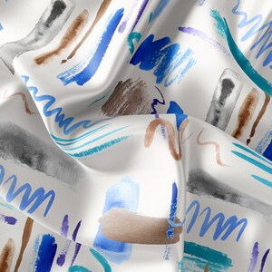 Op de afbeelding: Abstract waterverfpatroon op witte stof met blauwe, bruine en grijze penseelstreken. De stof is gevouwen en heeft textuur.