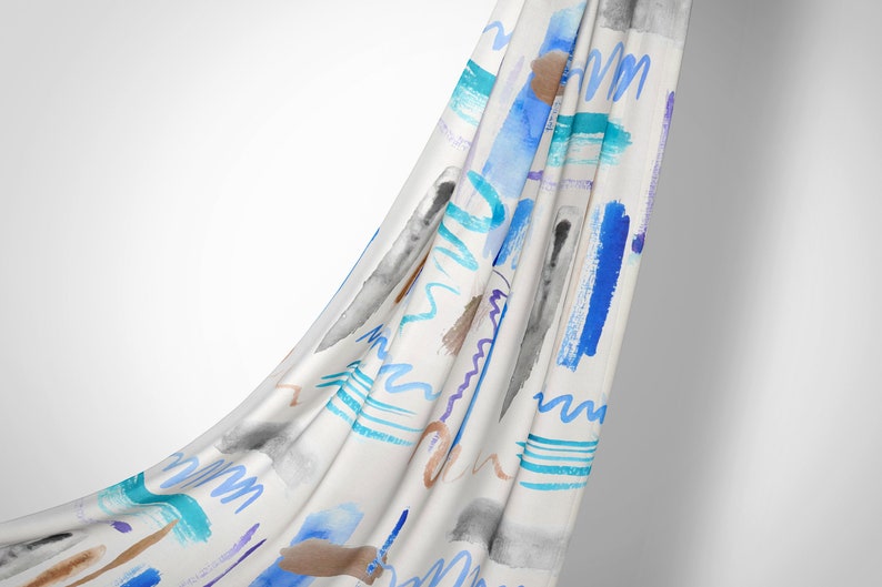 Op de afbeelding: Witte stof met een abstract aquarelpatroon in blauw, turquoise, paars en bruin. De stof is gedrapeerd en vloeit.