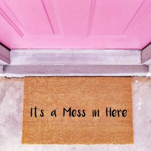 Puede incluir: Una alfombra de coco marrón con el texto "It's a Mess in Here" impreso en negro.