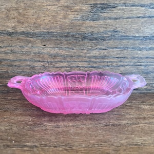 May include: A pink, oval-shaped glass dish with two small handles. The dish has a scalloped edge and a decorative pattern on the inside. The glass has an iridescent quality, reflecting light.