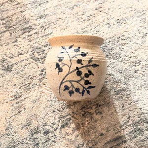 May include: A small, cream-colored ceramic vase with a blue floral design. The vase has a round body and a slightly flared lip.
