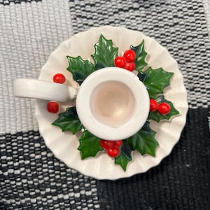 May include: White ceramic candle holder with a green holly leaf and red berry design. The holder has a scalloped edge and a round candle cup.