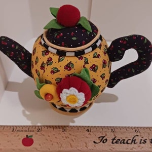 May include: A yellow and black teapot pincushion with a cherry print. The teapot has a red and white flower embellishment and a black handle. The pincushion is sitting on a wooden ruler with the text "To teach is to touch a life" written on it.