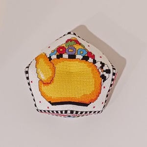 May include: A handmade, cross-stitched teapot design on a white, pentagon-shaped fabric. The teapot is yellow with a black and white checkered border, and a colorful floral design on top. The edges are trimmed with black and white stripes.