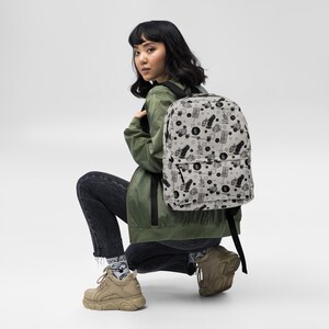 May include: A light gray backpack featuring an all-over black graphic print of various small illustrations, including figures, shapes, and symbols. The backpack has a large main compartment, a front zippered pocket, and black adjustable shoulder straps.