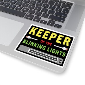 Puede incluir: Una pegatina negra y amarilla con el texto "KEEPER OF THE BLINKING LIGHTS" y un gráfico de un conmutador de red con cables Ethernet.