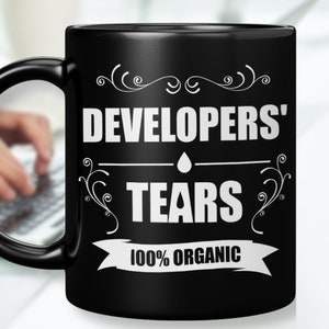 Peut inclure: Mug à café noir avec du texte blanc qui dit "Developers' Tears 100% Organic". Le texte est entouré de volutes décoratives.