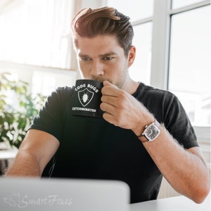 Pode incluir: Uma pessoa vestindo uma camiseta preta com uma caneca branca que diz "Code Bugs Exterminator" e um &iacute;cone de bug. A pessoa est&aacute; segurando a caneca na m&atilde;o direita e est&aacute; usando um rel&oacute;gio de couro marrom no pulso esquerdo.