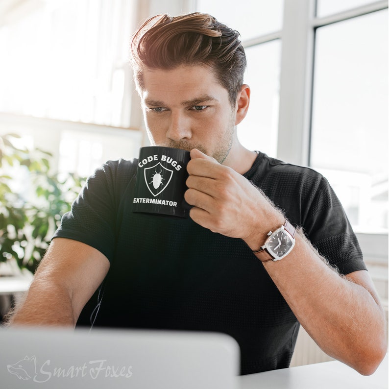 Pode incluir: Uma pessoa vestindo uma camiseta preta com uma caneca branca que diz "Code Bugs Exterminator" e um &iacute;cone de bug. A pessoa est&aacute; segurando a caneca na m&atilde;o direita e est&aacute; usando um rel&oacute;gio de couro marrom no pulso esquerdo.
