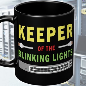 Puede incluir: Taza de cerámica negra con el texto "KEEPER OF THE BLINKING LIGHTS" en amarillo y verde. La taza presenta un gráfico de un conmutador de red con cables Ethernet.