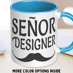 Könnte beinhalten: Weiße Keramik-Kaffeetasse mit blauem Rand und Henkel. Die Tasse hat einen schwarzen Schnurrbart und den Text "SEÑOR DESIGNER" in Schwarz.