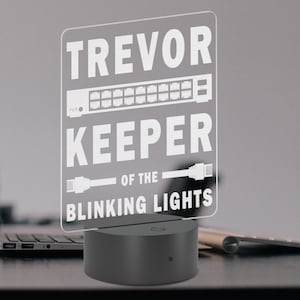 Könnte beinhalten: Ein weißes Acrylschild mit dem Text "Trevor Keeper of the Blinking Lights" und einer Grafik eines Netzwerk-Switches und Ethernet-Kabeln. Das Schild wird von hinten beleuchtet und steht auf einem schwarzen Sockel.