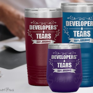 以下が含まれることがあります： 紫、赤、青のステンレス製タンブラー3個。各タンブラーには、「Developers' Tears 100% Organic」の文字とスタイリッシュなデザインが施されています。