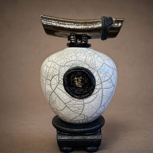 May include: A ceramic sculpture featuring a white, cracked-glaze vase with a black base and a dark, curved top element. A black seal with gold text is affixed to the vase. The sculpture has a modern, artistic aesthetic.