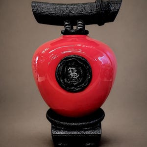 May include: A red, glossy decorative vase with a black, textured top and base. The vase has a circular black emblem with an Asian character. The vase is a unique decorative item.