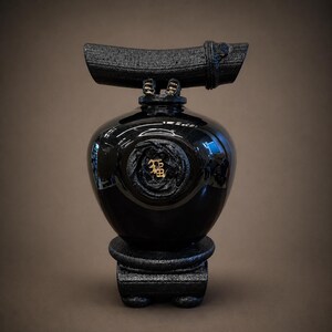May include: A black decorative vase with a glossy finish, featuring a unique design. The vase has a rounded body, a textured top, and a pedestal base. A gold emblem is centered on the vase. The overall aesthetic is modern and elegant.