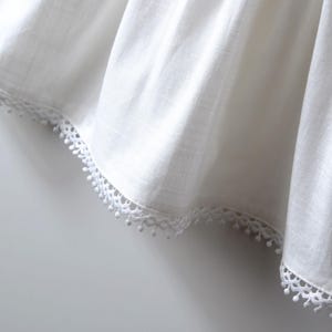 May include: A white fabric garment featuring a subtle vertical weave texture and a decorative scalloped trim along the bottom edge. The trim consists of a delicate geometric pattern accented with small, evenly spaced hanging pom-pom details.