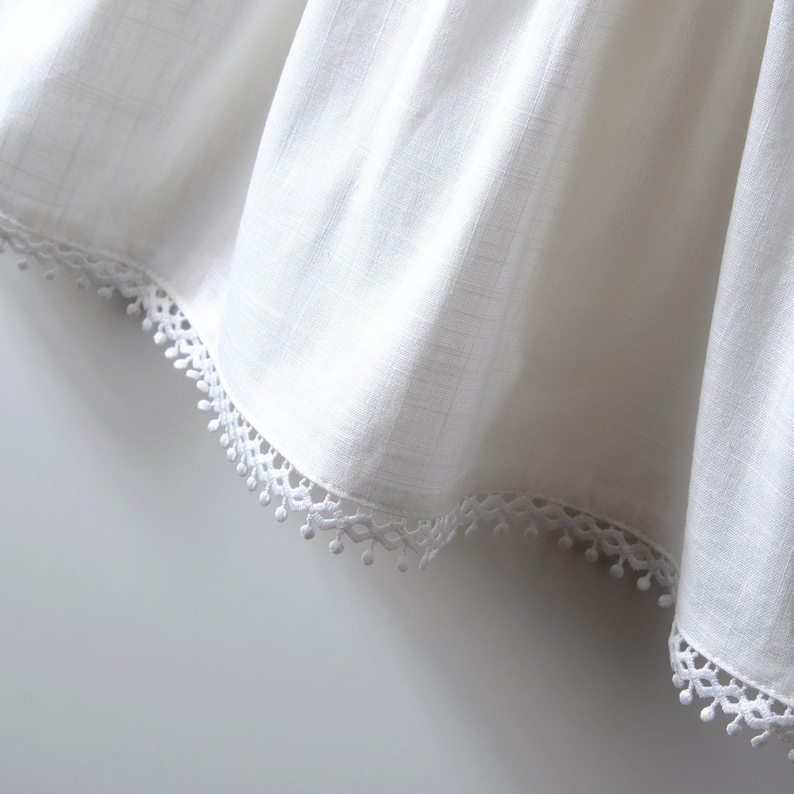 May include: A white fabric garment featuring a subtle vertical weave texture and a decorative scalloped trim along the bottom edge. The trim consists of a delicate geometric pattern accented with small, evenly spaced hanging pom-pom details.