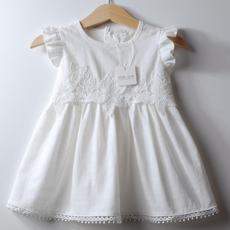 May include: A white sleeveless dress featuring ruffled cap sleeves, a round neckline, and a floral lace applique across the chest. The hem is finished with a delicate scalloped lace trim. A tag labeled Studio Loliwe is attached to the neckline.