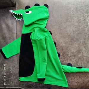 Può includere: Costume da dinosauro verde con cappuccio, dettagli neri e coda. Il costume ha una cerniera e un cappuccio con la faccia di un dinosauro.