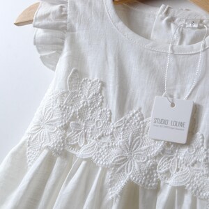 May include: A white sleeveless dress featuring ruffled cap sleeves and a prominent floral lace applique across the chest. The garment includes a white tag with the text STUDIO LOLIWE and a size 6 label attached to the neckline area.