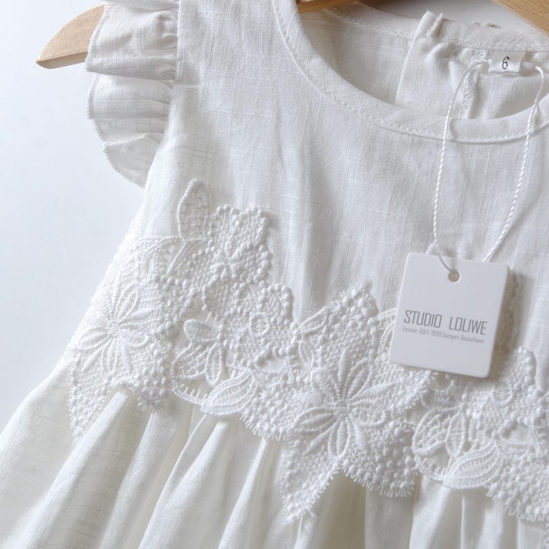 May include: A white sleeveless dress featuring ruffled cap sleeves and a prominent floral lace applique across the chest. The garment includes a white tag with the text STUDIO LOLIWE and a size 6 label attached to the neckline area.