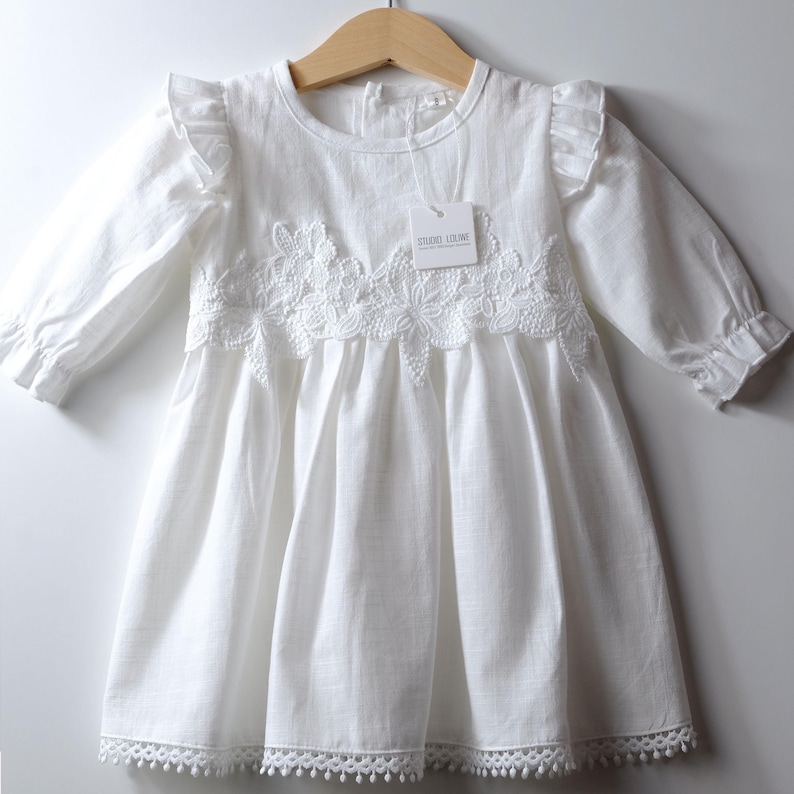 May include: A white long-sleeved dress featuring a round neckline, ruffled shoulders, and a gathered skirt. The bodice is adorned with a white floral lace appliqu&eacute; across the chest. A white tag labeled STUDIO LOLIWE is attached to the neckline.
