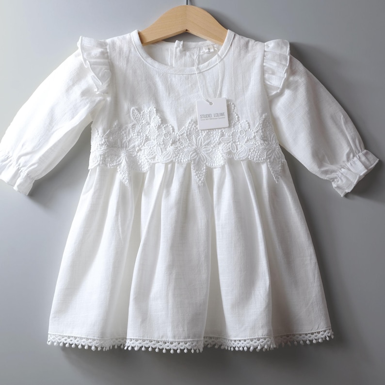 May include: A white long-sleeved dress featuring ruffled shoulders, a round neckline, and a gathered skirt. The bodice is adorned with white floral lace embroidery. A small white tag labeled STUDIO LOUWE is attached to the neckline.
