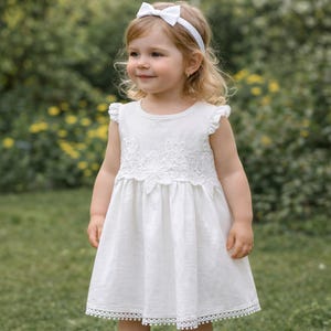 May include: A white sleeveless dress featuring a round neckline, ruffled shoulders, and a floral lace applique across the chest. The hem is finished with a delicate pom-pom trim. A matching white headband with a bow is worn on the head.