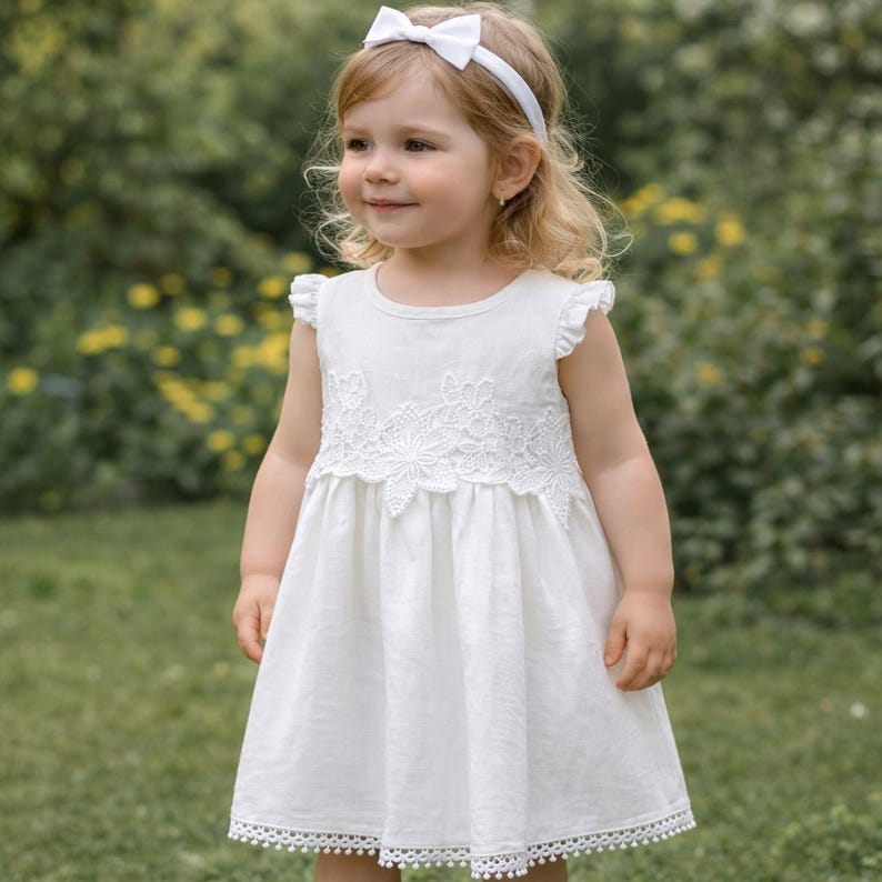 May include: A white sleeveless dress featuring a round neckline, ruffled shoulders, and a floral lace applique across the chest. The hem is finished with a delicate pom-pom trim. A matching white headband with a bow is worn on the head.