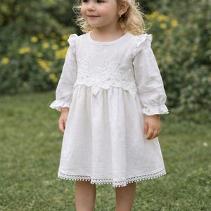 May include: A white long-sleeved dress featuring ruffled shoulders, a lace bodice overlay, and a pom-pom trim hem. The outfit includes a matching white floral lace headband and white shoes adorned with cream-colored bows and small crystal accents.