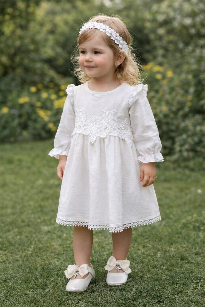 May include: A white long-sleeved dress featuring ruffled shoulders, a lace bodice overlay, and a pom-pom trim hem. The outfit includes a matching white floral lace headband and white shoes adorned with cream-colored bows and small crystal accents.