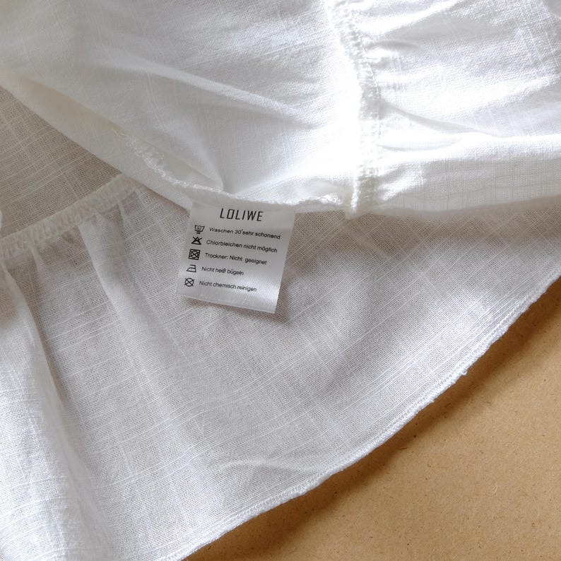 May include: A white garment with a subtle grid texture and a ruffled hem. A white care label is attached to the interior, featuring the brand name LOLIWE and German care instructions including symbols for washing, bleaching, drying, and ironing.